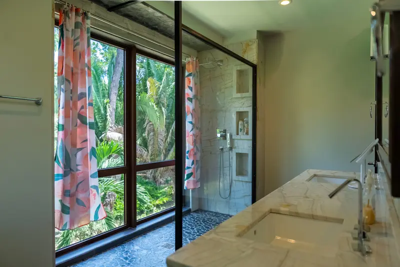 Walk-in shower with marble walls, glass screen and floral curtain beside palm-view window