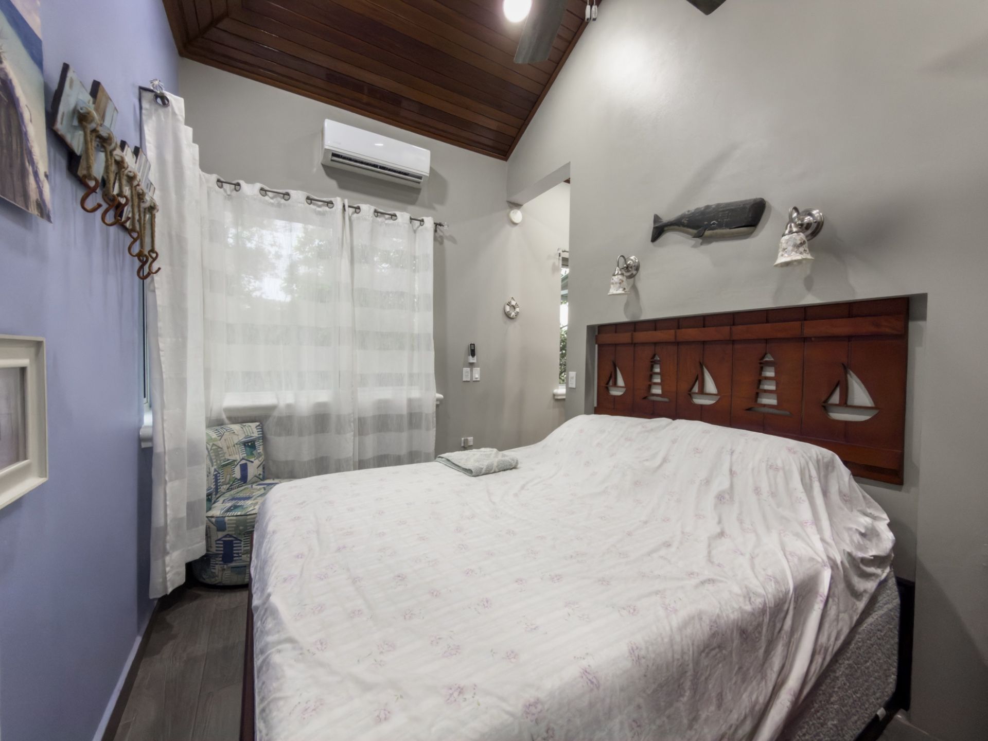 Bedroom with white bed, wooden headboard with sailboat carvings and wood-paneled ceiling