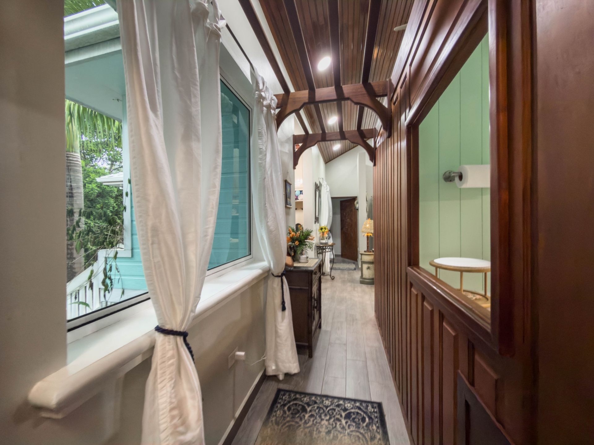 Narrow hallway with wood-paneled ceiling, white curtains and potted flowers on a dark console