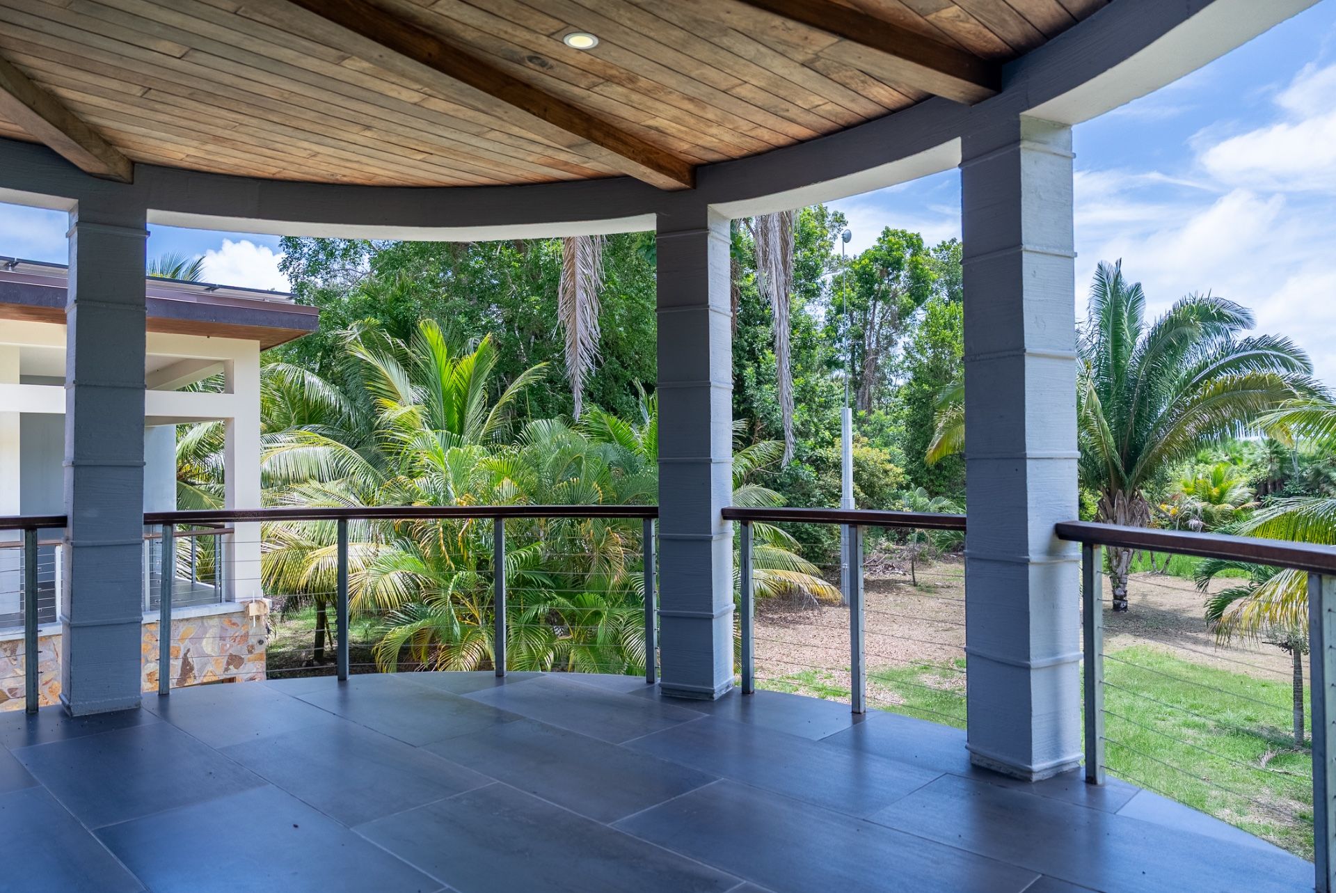Covered deck with dark tile floor, wood ceiling and cable railing over palm trees