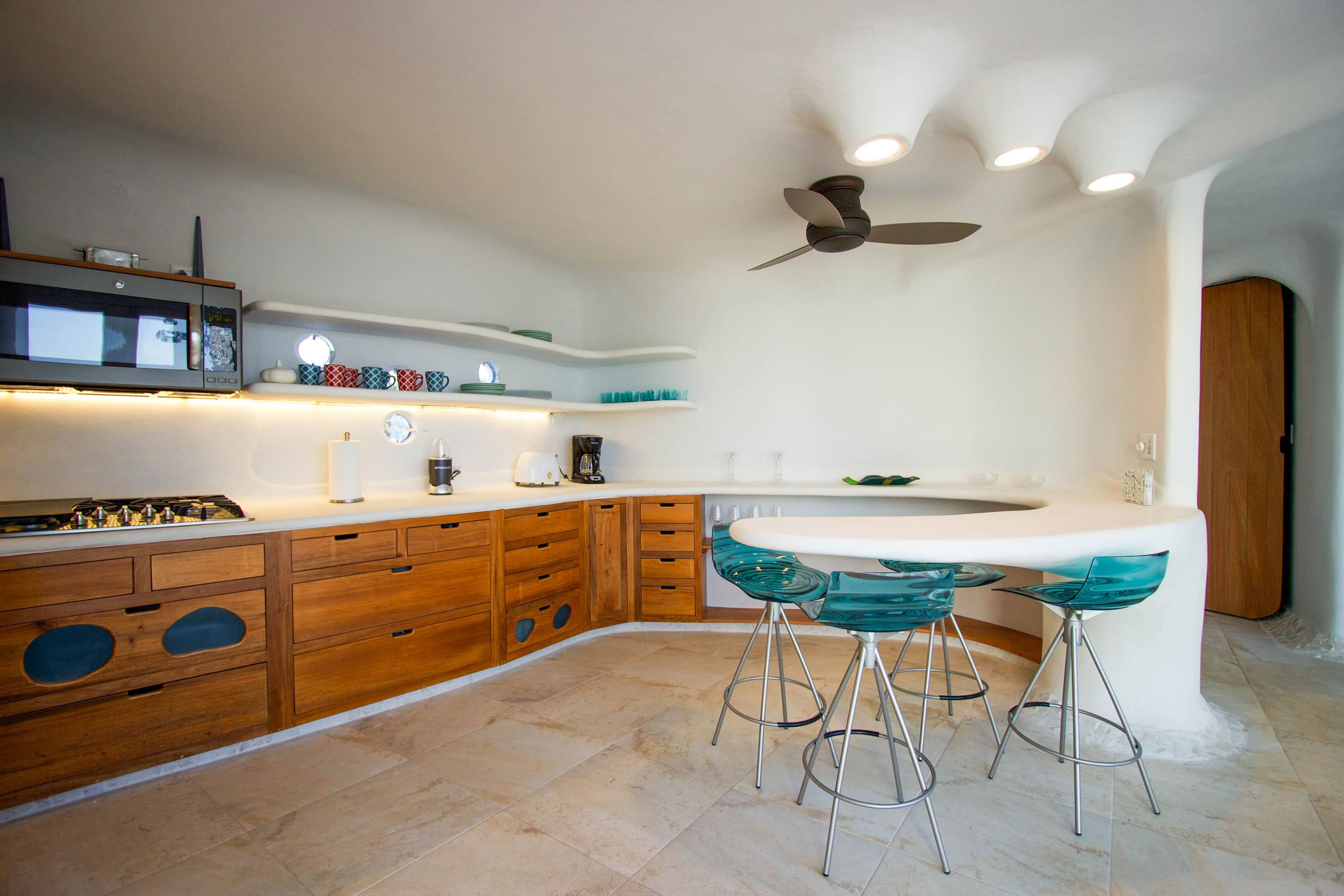 Compact kitchen with wooden cabinets, white curved shelves and teal glass stools