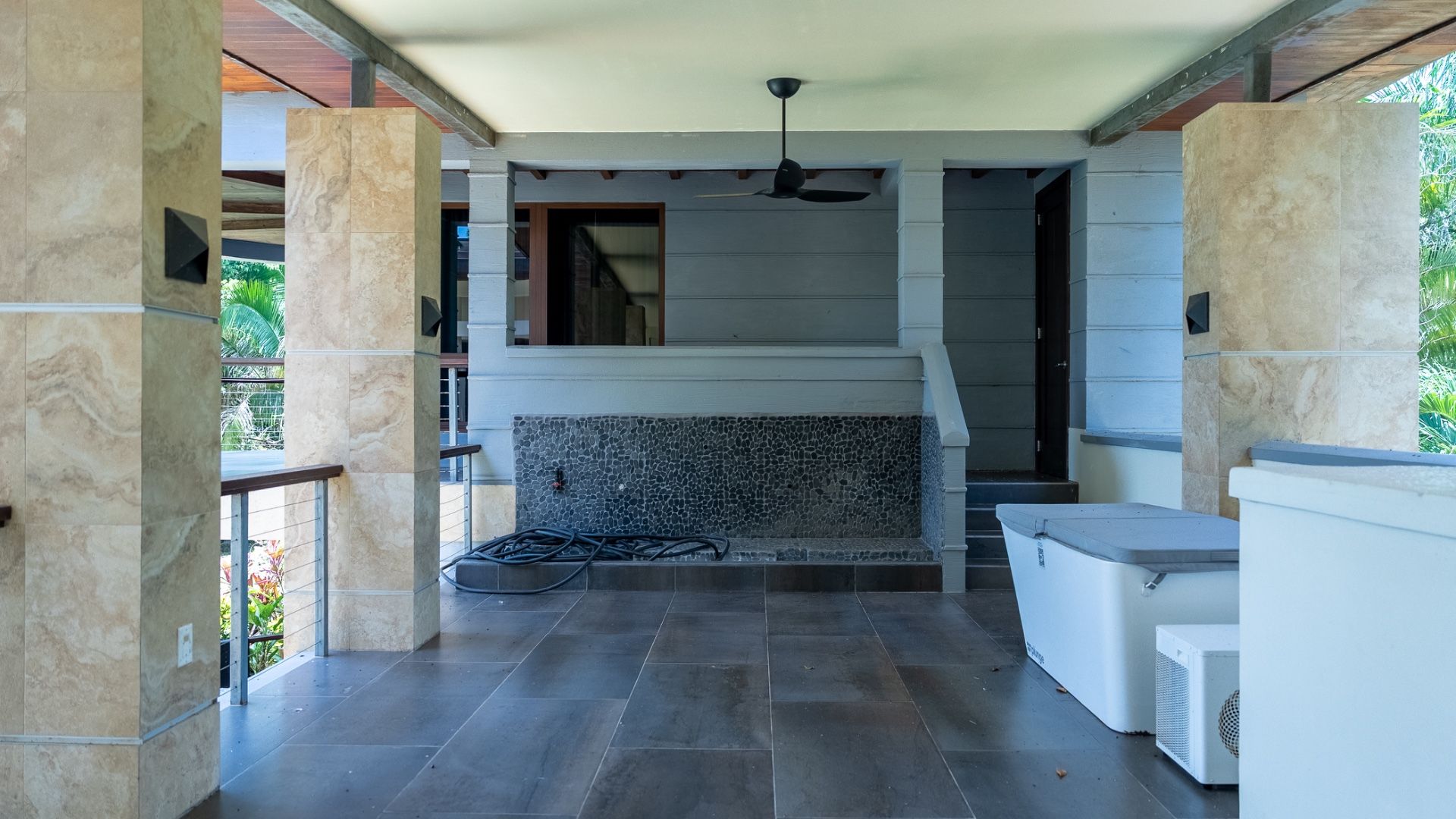 Covered porch with dark tile floor, black ceiling fan and pebble-tiled wall