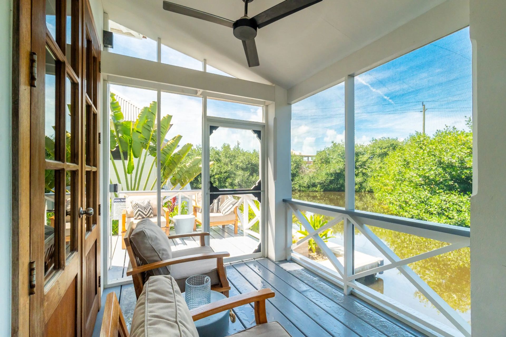 Screened porch with wooden lounge chairs, ceiling fan and water view beyond the railing