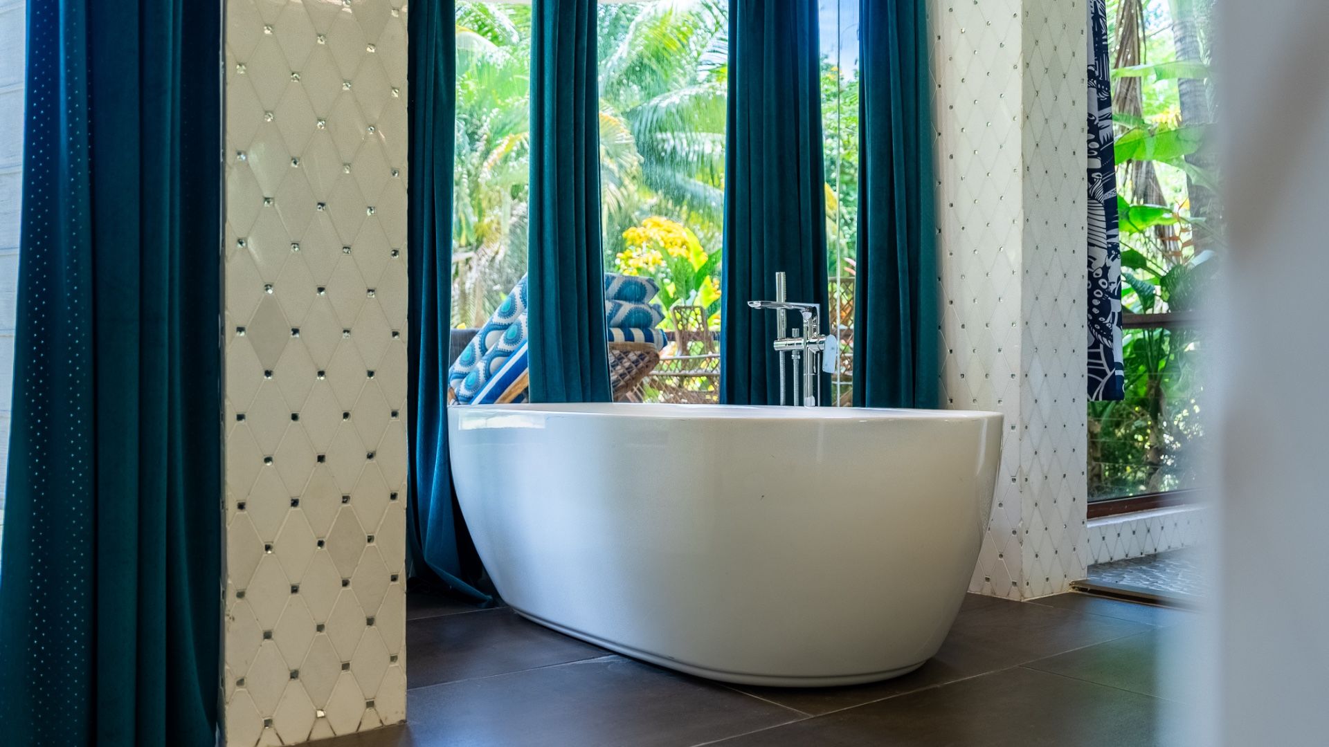 Freestanding white bathtub before teal curtains and a view of tropical garden