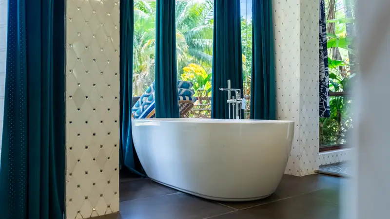 Freestanding white bathtub before teal curtains and a view of tropical garden