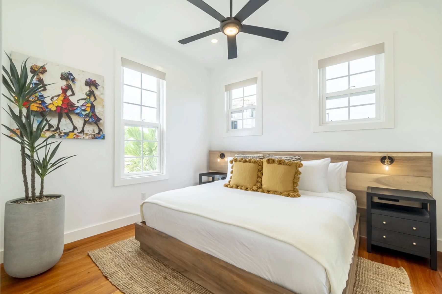 Bedroom with white bed, wood frame and mustard tassel pillows under a ceiling fan